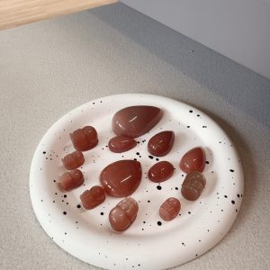 A plate of sunstone rough stones in various shapes, including teardrop and carved pieces, with a warm reddish-brown color, placed on a white disc with black spots, against a background of light gray and wooden floor, showcasing the diverse shapes and textures of sunstone rough stones.