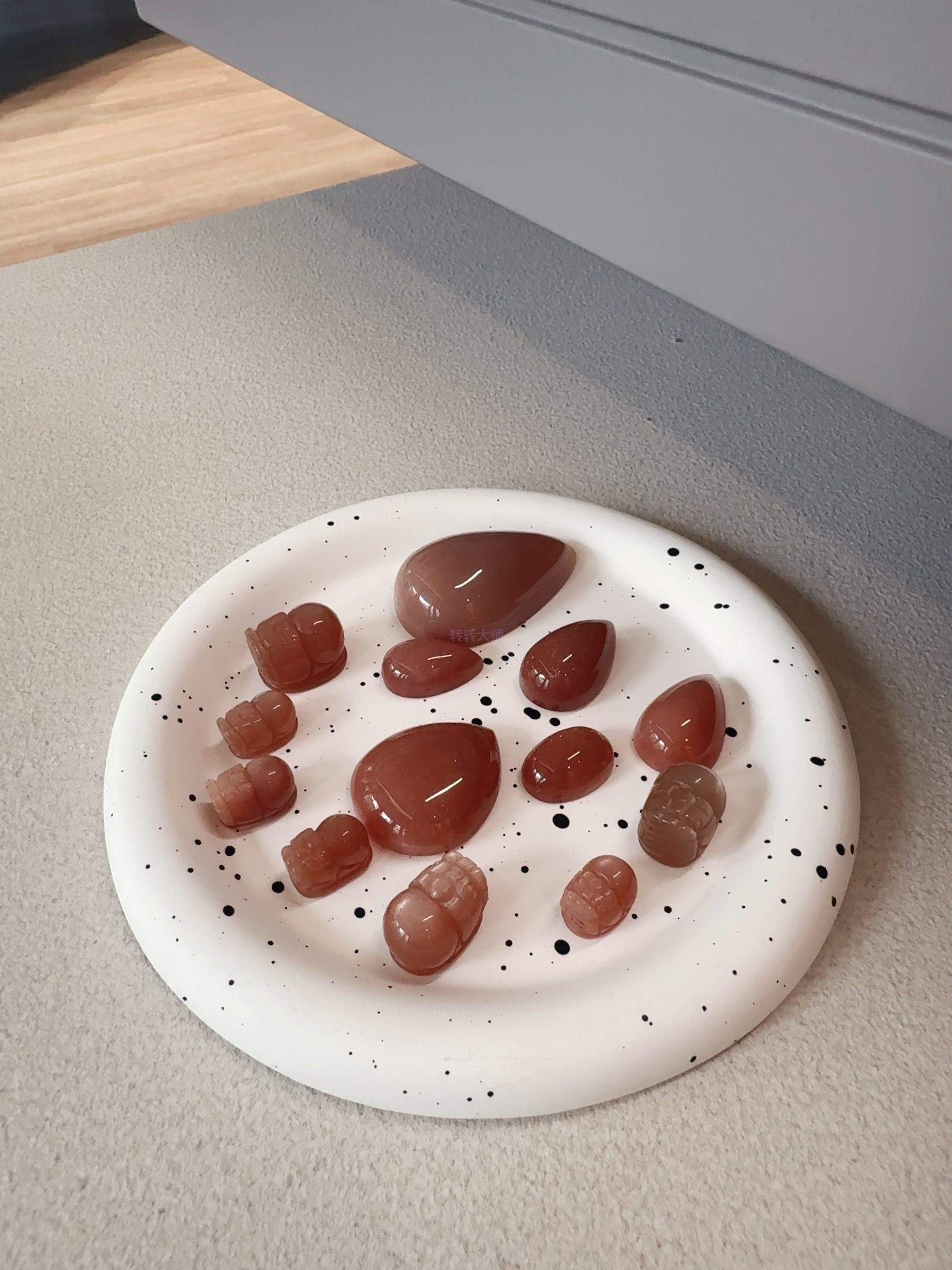 A plate of sunstone rough stones in various shapes, including teardrop and carved pieces, with a warm reddish-brown color, placed on a white disc with black spots, against a background of light gray and wooden floor, showcasing the diverse shapes and textures of sunstone rough stones.