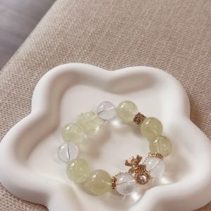 A bracelet made of lemon crystal and white crystal, paired with a gold diamond-studded money bag accessory, placed on a white flower-shaped tray with a light beige fabric background, presenting the fresh and elegant texture and design details of crystal jewelry.
