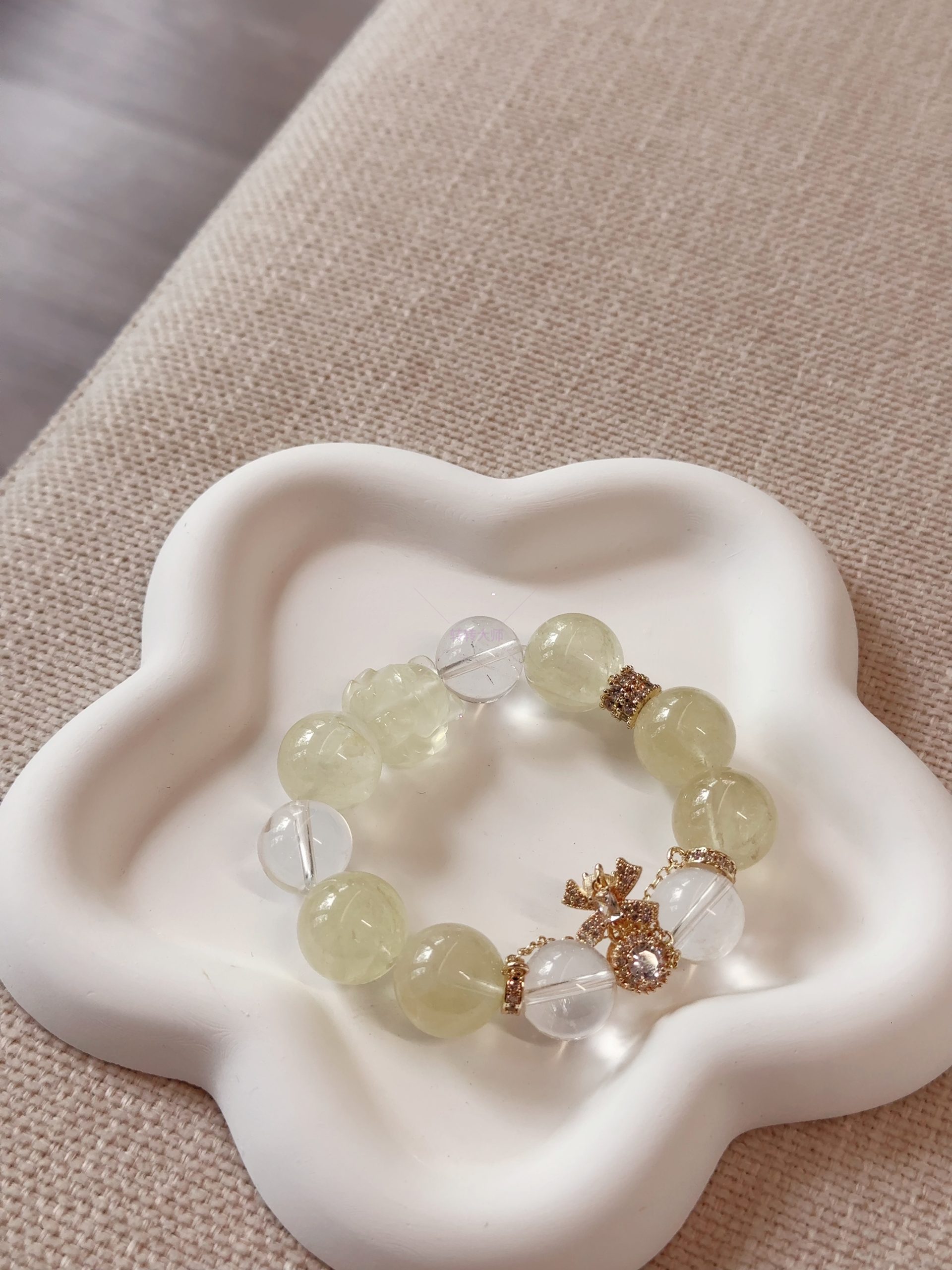 A bracelet made of lemon crystal and white crystal, paired with a gold diamond-studded money bag accessory, placed on a white flower-shaped tray with a light beige fabric background, presenting the fresh and elegant texture and design details of crystal jewelry.